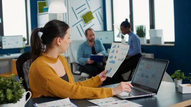 Woman comparing statistics with sales charts on laptop and taking a break to drink coffee or tea sitting at desk. Casual startup employee working relaxed in busy finance department analyzing
