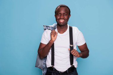 Handsome and confident looking guy posing for camera while wearing suspenders and tshirt. Cheerful young adult man having denim jacket while smiling at camera and standing on blue background.