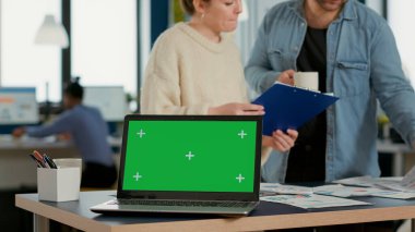 Laptop computer with green screen on desk in front of startup colleagues talking about sales charts papers on clipboard. Focus on portable computer screen with chroma key empty background.