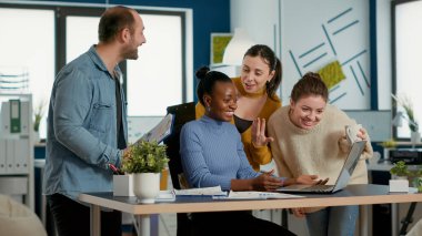 Smiling successful employee with great work results receiving congratulations from coworkers at desk. Woman working on laptop happy with business turnover celebrates with team in startup office.