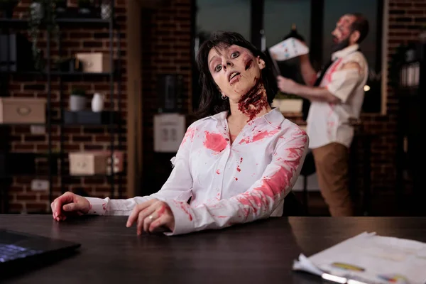 Portrait of scary corpse with wounds in office, sitting at desk to work on laptop being terrifying and spooky. Cruel possessed brain eating monster with bloody mouth open, walking dead devil.