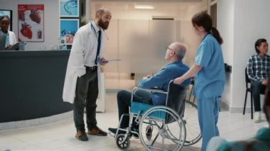 Male doctor meeting with old patient in wheelchair at reception lobby, having conversation about healthcare and support. Man with chronic disability having checkup visit with specialist.