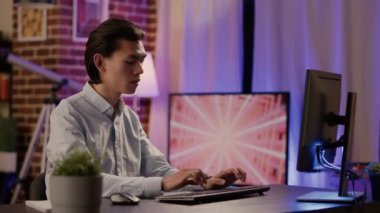 Asian male freelancer browsing internet website on computer, writing email and finance report to create business presentation. Working from home on pc, doing paperwork. Tripod shot.