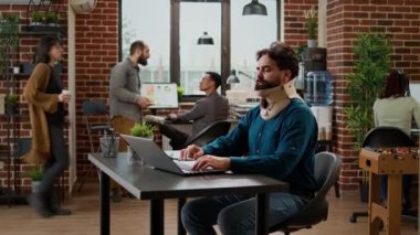 Business man in pain wearing cervical neck foam at office job, working on laptop and being hurt. Male employee with medical collar brace suffering after physical accident injury at work.