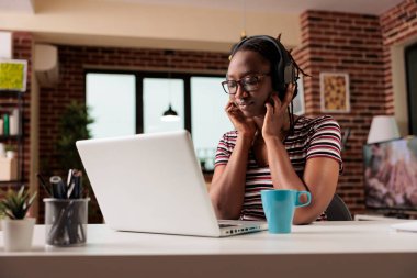 Young african american woman in wireless headphones watching movie online on streaming service on laptop, freelancer having break from work. Teenager leisure activity, home entertainment