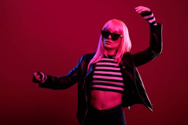 Caucasian trendy person with big sunglasses and leather jacket feeling beautiful and confident posing on camera. Standing in studio with stylish fashion items to present amazing style in dark light.