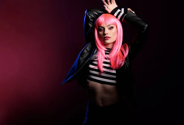 Portrait of sensual beauty model with pink hair wig and rocker jacket standing over background with dark shadows and light. Sexy stylish woman with punk trendy fashion items doing pose.