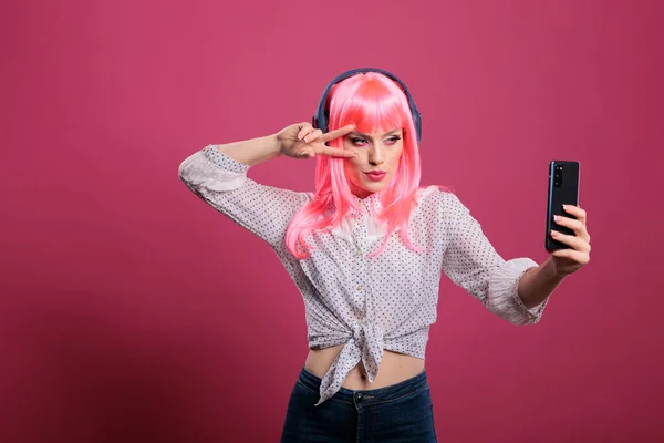 Modern cool woman taking pictures with smartphone app and listening to music, standing over pink background. Using mobile phone to have fun with camera photos, cheerful happy model.