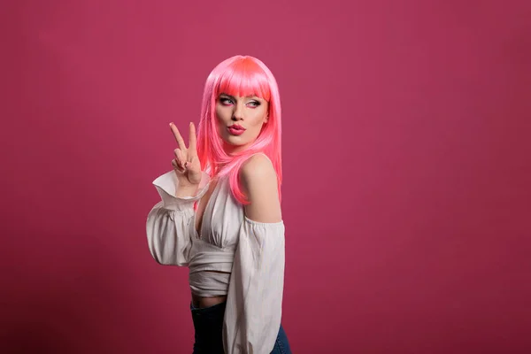 Trendy cool woman with pink hair doing peace sign and kissy face in front of camera. Carefree attractive girl showing victory symbol and peaceful gesture with two fingers up in studio.