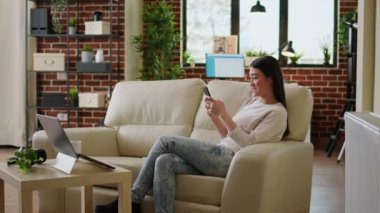 Smiling heartily asian woman watching funny content on smartphone while drinking coffee at home. Joyful young adult person texting with friend while sitting on sofa inside apartment.