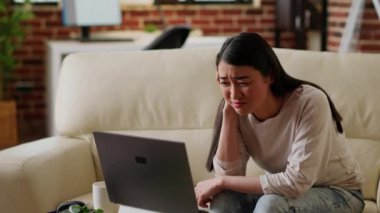 İş ödevini anlamaya çalışırken dizüstü bilgisayarda çalışan stresli bir kadın. Endişeli Asyalı kişi uzaktan kumandayla uğraşırken bilgisayar ekranına şaşkın bakıyor.