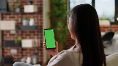 Asian woman having smartphone with green screen display while sitting at home on sofa. Young adult person sitting on couch with mobile cellphone having mockup chroma key background.