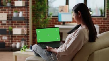 Young woman sitting at home with laptop having green screen chroma key display. Adult person sitting on sofa with portable computer on lap having mockup isolated template background. Tripod shot