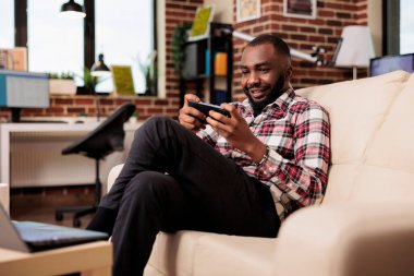 Freelancer playing online gaming competition on smartphone, having fun with video games and sitting on couch. Enjoying remote internet game play on mobile phone app, working on freelance job.