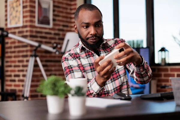 African american man playing video games on mobile phone, having fun with online gaming competition and working from home. Enjoying game play on smartphone, freelaner doing leisure activity.