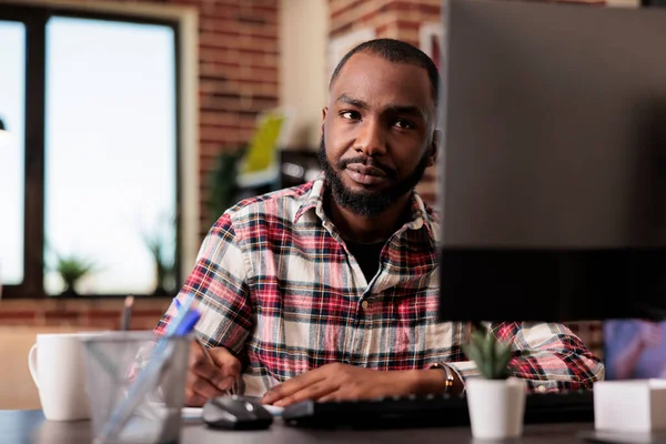 African american man taking notes on files, writing report information on notebook at office desk. Working from home on startup documents, doing paperwork and browsing internet on computer.