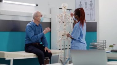 Medical assistant showing human skeleton bones to old patient at consultation during covid 19 pandemic. Nurse and senior man analyzing anatomy spinal cord to find orthopedic diagnosis.