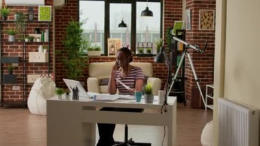 African american woman talking on mobile phone call at home, doing remote work on laptop. Calling people on smartphone to discuss on telephone line, telework job and telecommunications.