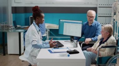 Health specialist consulting senior woman with impairment, explaining diagnosis to wheelchair user and husband at medical appointment. Doctor examining old patient with chronic disability.
