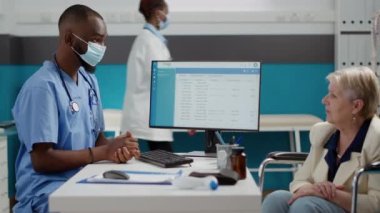 Specialist consulting old woman with disability in office, having medical appointment at facility. Patient in wheelchair talking to nurse with face mask about disease and impairment.