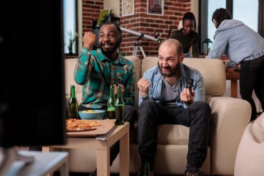 Cheerful people celebrating video games win at house gathering, using tv console to play competition. Men feeling happy about winning, having fun wiht friends at party with beer.