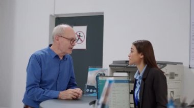 Senior man asking for help at reception desk in facility lobby, talking to female receptionist about healthcare insurance and appointment. Old patient doing checkup consultation at health center.