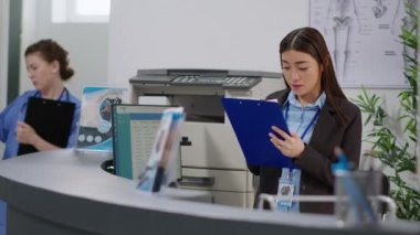 Woman receptionist working on healthcare appointments with checkup report papers at hospital reception desk. Using consultation forms to give insurance support and help with examinations at counter.