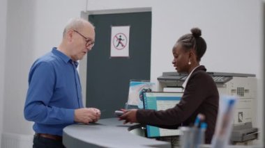 Old man using credit card to pay after consultation appointment, making payment transaction at hospital reception counter. Senior patent paying for checkup visit and treatment.
