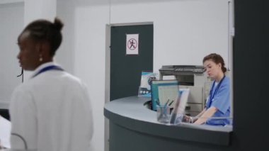 Medical assistant working at hospital registration counter with consultation appointments on computer. Helping with checkup visits and health insurance at facility reception desk. Handheld shot.