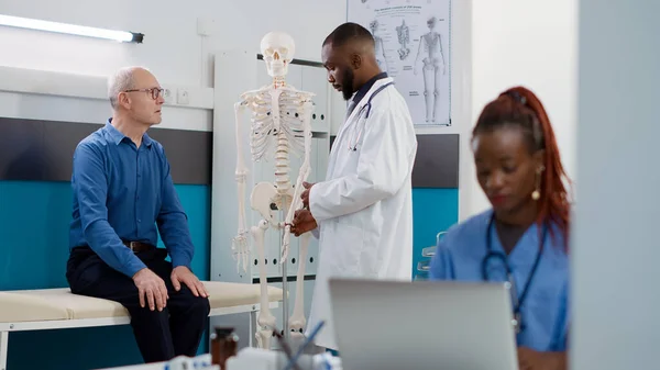 非裔美国人骨科病患者指向人体骨骼 向老年患者解释骨病和诊断 并进行检查 骨病检查显示脊髓的解剖学专家 — 图库照片