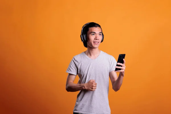Asian man in wireless headphones enjoying music, smiling teenager listening to educational podcast. Young person standing in earphones, holding smartphone, studio mid shot
