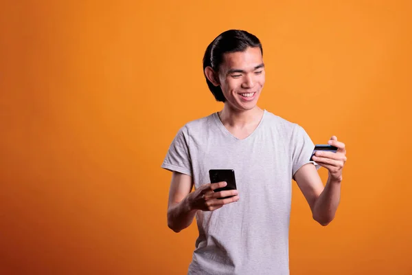 Smiling young man buying products in internet shop, making order using smartphone app. Asian teenager holding debit card and searching goods in online store, e commerce concept