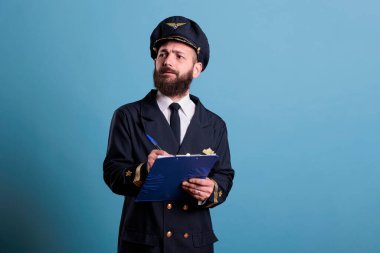 Uçak kaptanı pano tutuyor, evrakları dolduruyor, yoğunlaştırılmış havacı formunu dolduruyor. Havacılık üniformalı bir uçak pilotu, hava alanında ön planda yazıyor, mavi arkaplanda stüdyo resmi var.