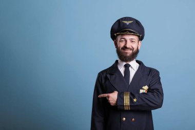 Uçak pilotu işaret parmağıyla yan tarafı işaret ediyor, reklam ürünü, profesyonel üniforma giyiyor. Gülümseyen uçak kaptanı kameraya bakıyor, terfi jesti yapıyor.