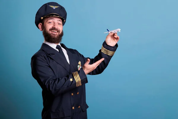 Smiling pilot with uniform playing with airplane model side view, aviation academy aviator holding commercial plane toy. Funny aircraft capitan, studio medium shot on blue background