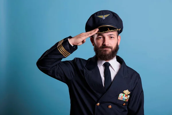 Confident airplane pilot saluting, wearing uniform and hat front view portrait, plane captain looking at camera. Civil aviator with airline wings badge on jacket, professional aircrew member