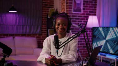Afro-Amerikan Podcaster 'ın portresi evde kayıt stüdyosunda mikrofon ve dijital ses mikseriyle oturuyor. Çevrimiçi radyo sunucusu canlı yayın yaparken kameraya kendinden emin bir şekilde gülümsüyor.