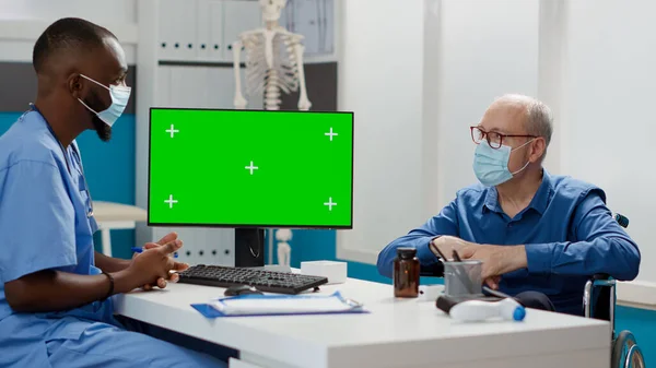 Nurse and senior man with disability looking at greenscreen on monitor in cabinet. Wheelchair user and specialist with face masks using isolated chroma key display with mockup copyspace.