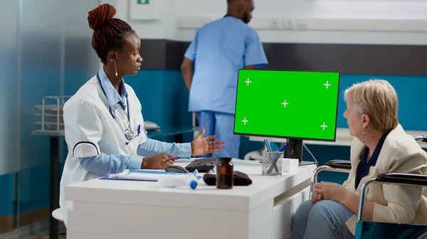 Specialist pointing at computer with greenscreen display at checkup with old woman in wheelchair. Medic and patient looking at chroma key background with isolated mockup copyspace.