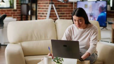 Focused student taking notes while attending online class on laptop at home. Young adult woman writing on notebook while being attentive to modern portable computer in living room.