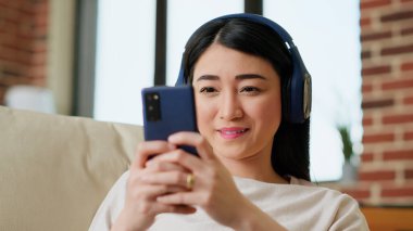 Cheerful girl texting friend on touchscreen phone while working remotely. Cheerful woman sending messages on mobile phone while listening to music and doing work from home. Handheld shot