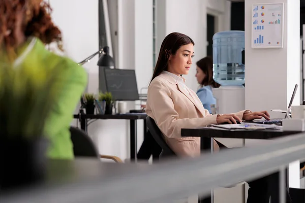 Young asian woman working on laptop in coworking space, busy company worker sitting at workplace desktop. Serious employee using computer in modern corporate office, side view