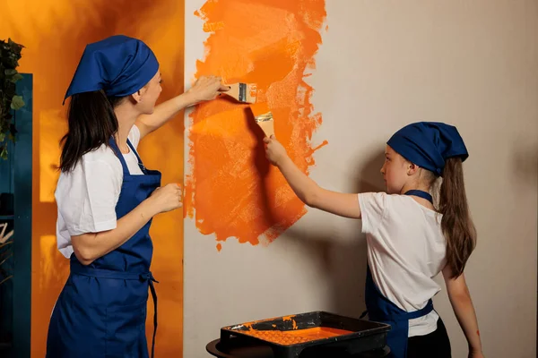 Mom and kid using orange paint on walls and brush to renovate house interior, painting apartment room together. People having fun with housework redecoration, diy decorating job.