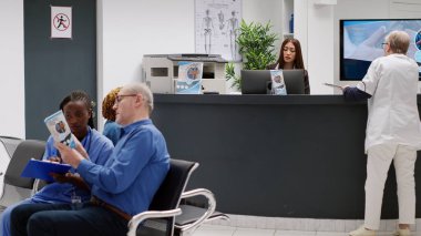 Female receptionist talking to senior doctor at reception counter in medical facility waiting area. General practitioner asking worker about medical reports and healthcare appointments.