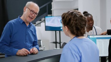 Medical assistant helping senior patient with appointment and reception registration counter in healthcare facility. Nurse working at hospital help desk in lobby. Handheld shot.