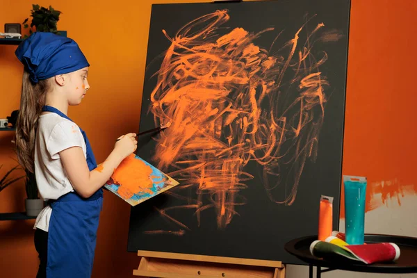 Small girl using orange paint color on canvas, creating artwork masterpiece with colormix tray and aquarelle palette. Painting inspiration design with painbrush, watercolor and skills.
