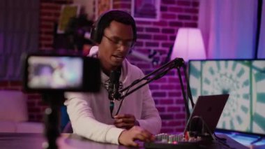 Online radio host answering internet call from listener while recording vlog using digital video camera in home podcasting studio. African american podcaster talking to audience using microphone.