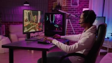 African american gamer playing intense action game in tournament talking with team using gaming headset at home. Man having intense experience streaming online first person shooter game on pc station.