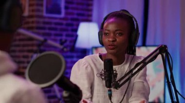 Vlogger 'ın masasında oturduğu intervew' deki ev kayıt stüdyosundaki Afro-Amerikan podcaster 'a yakın çekim. Çevrimiçi radyo sunucusu, mikrofonla canlı yayın yaparken kameraya kendinden emin bir şekilde gülümsüyor..