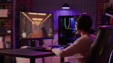 Gamer girl using pc setup playing multiplayer first person shooter talking on headset while explaining gameplay to subscribers. Woman streaming online action game while sitting in gaming chair.